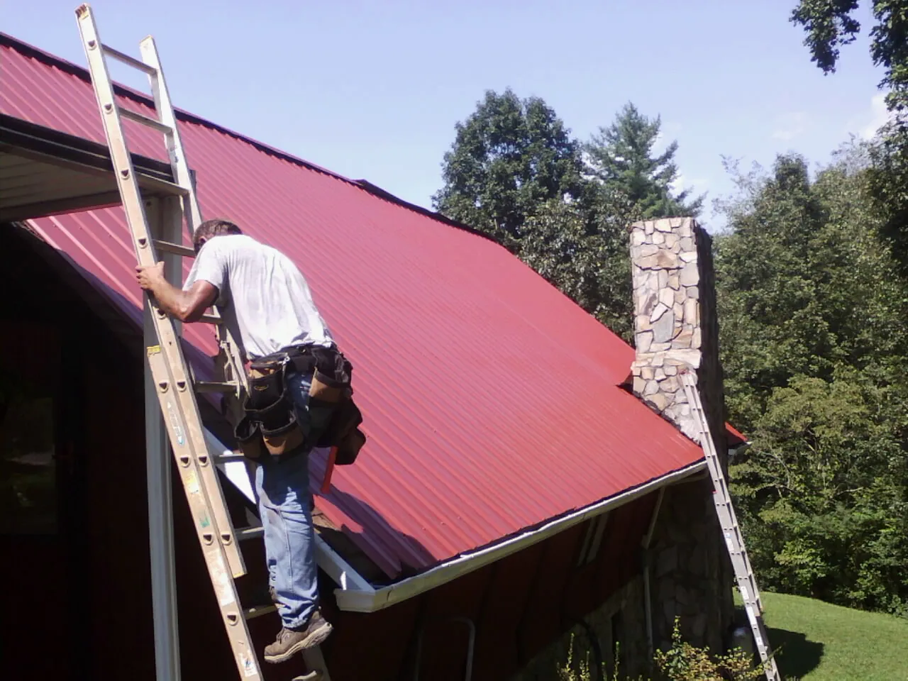 Roofer on a ladder beside a red metal roof and stone chimney — Soffit & Fascia Repair by Roofing Brothers in Cave City